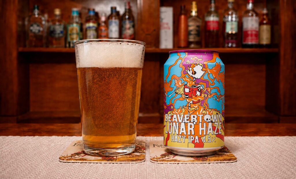 Glass of beer with foamy head beside a vibrant Beavertown Lunar Haze IPA can on a bar counter.